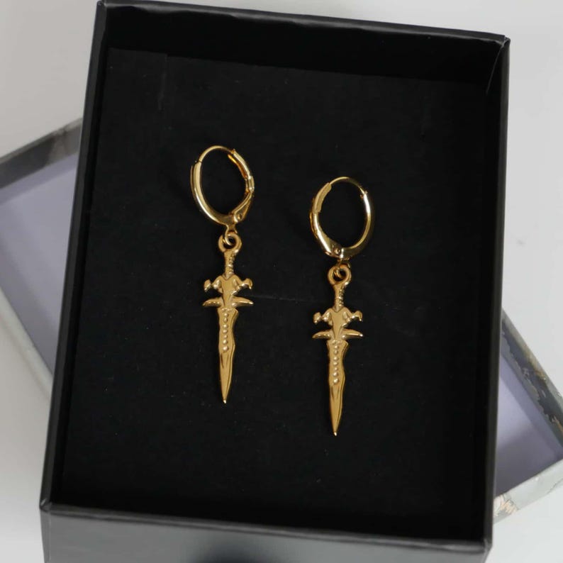 May include: A pair of gold-toned hoop earrings featuring dangling sword-shaped charms. Each earring consists of a circular hoop connected to a detailed, pointed blade pendant with a crossguard. The jewellery is presented inside a black box.