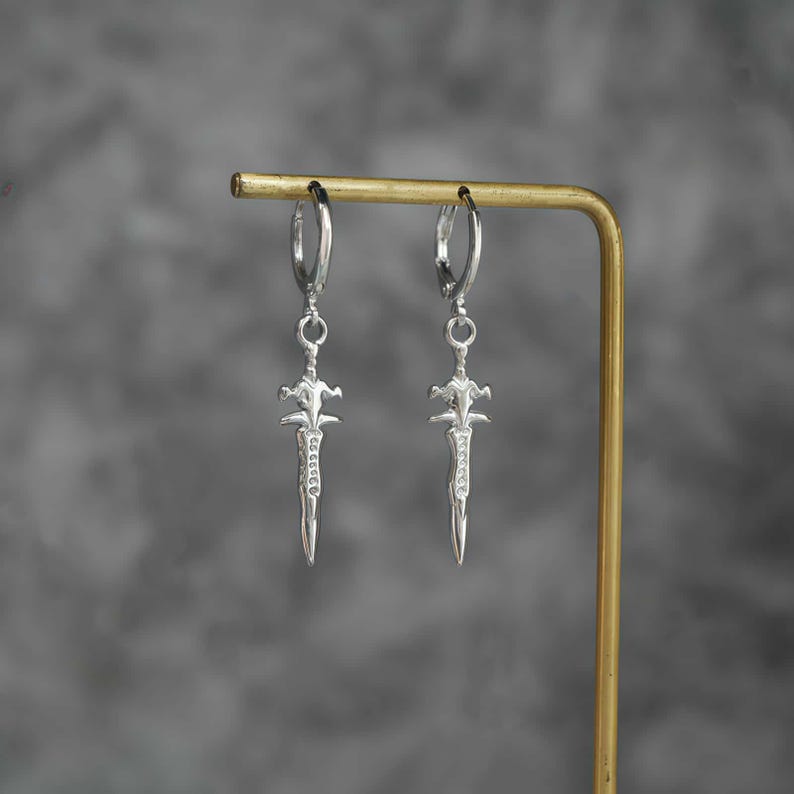 May include: A pair of silver-toned hoop earrings featuring a dangling sword-shaped charm. Each charm has a pointed blade, a crossguard, and a textured handle design. The earrings are suspended from a thin, circular hoop mechanism that secures with a hinge.