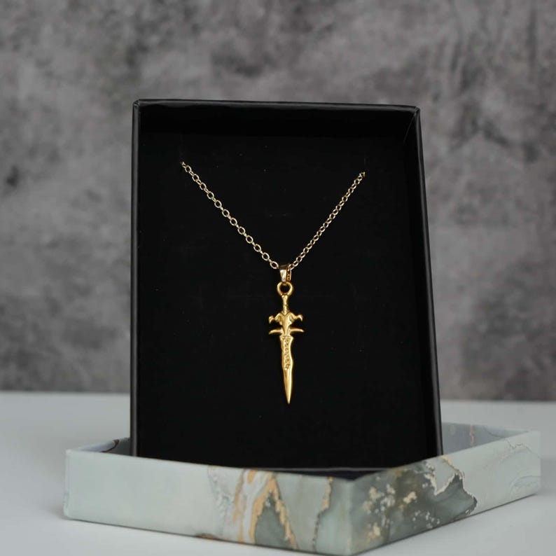 May include: A gold-coloured necklace featuring a pendant shaped like a small sword or dagger. The pendant hangs from a delicate matching gold-coloured chain. The jewellery is displayed against a black velvet-lined insert inside a rectangular box.