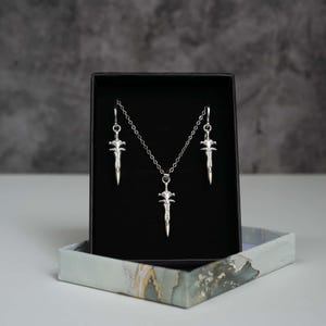 May include: A silver-toned jewellery set consisting of a necklace and two matching earrings, each featuring a small sword-shaped pendant. The pieces are displayed against a black velvet insert inside a rectangular box with a marble-patterned base.