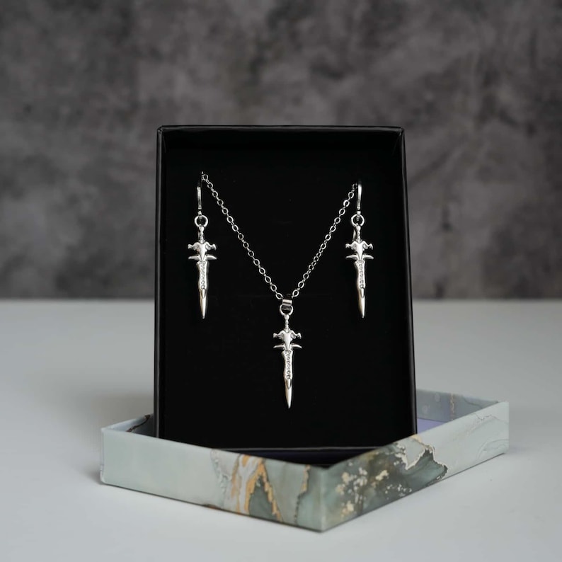 May include: A silver-toned jewellery set consisting of a necklace and two matching earrings, each featuring a small sword-shaped pendant. The pieces are displayed against a black velvet insert inside a rectangular box with a marble-patterned base.