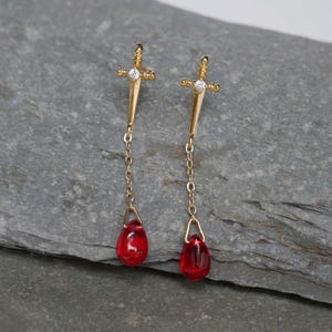 May include: Gold dangle earrings with a sword charm and a red teardrop-shaped glass bead at the end of each earring.