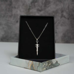 May include: A silver-toned necklace featuring a pendant shaped like a sword with a pointed blade and a crossguard. The pendant hangs from a delicate silver-toned chain, displayed against a black velvet-lined insert inside a rectangular box.