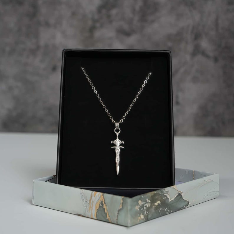 May include: A silver-toned necklace featuring a pendant shaped like a sword with a pointed blade and a crossguard. The pendant hangs from a delicate silver-toned chain, displayed against a black velvet-lined insert inside a rectangular box.