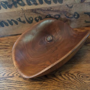 May include: A hand-carved wooden bowl with a natural, organic shape. The bowl is made of a rich, dark brown wood with visible grain patterns and a smooth, polished finish. The bowl has a shallow, curved design, perfect for displaying fruit or decorative items.