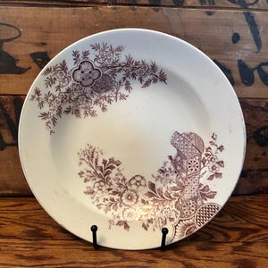May include: A white ceramic plate with a maroon floral pattern. The pattern includes flowers, leaves, and geometric shapes. The plate is displayed on a black metal stand against a wooden background.