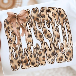 May include: White t-shirt with the word "BLESSED" in a leopard print design. A brown bow is attached to the upper left side of the text. The shirt is held by a person.