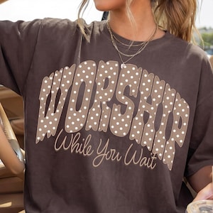 以下が含まれることがあります： 茶色のTシャツ。 「WORSHIP」の文字がドット柄で、「While You Wait」のフレーズが筆記体で書かれています。 車の中で人が着ています。