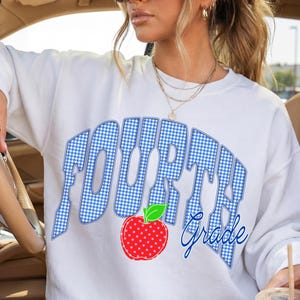 May include: White sweatshirt with the words "FOURTH Grade" in blue gingham lettering and a red apple graphic. The sweatshirt is a casual style, suitable for school or everyday wear.