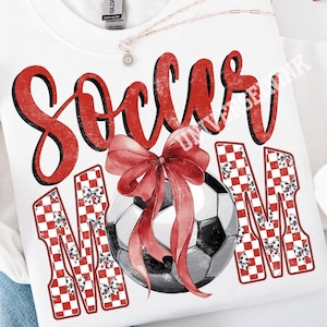May include: White sweatshirt with the words "Soccer Mom" in red script and block letters. A soccer ball is centered below the word "Soccer" and a red bow is on top of the soccer ball. The letters "MOM" are in a red and white checkered pattern.