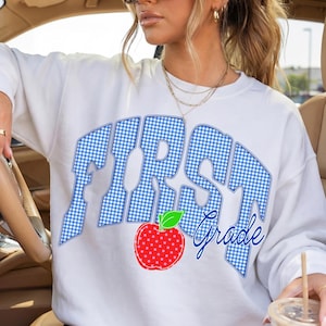 May include: White sweatshirt featuring the text "FIRST Grade" in a blue gingham pattern. A red apple with a green leaf is below the text. The sweatshirt is worn by a person in a car.