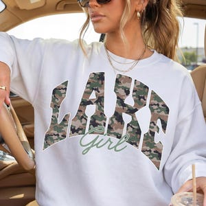 May include: White sweatshirt with the text "LAKE Girl" in a camouflage design. The sweatshirt has a crew neck and long sleeves. The text "Girl" is in a cursive font. The person is wearing sunglasses and rings.