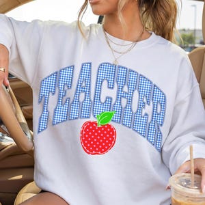 May include: White sweatshirt with the word "TEACHER" in blue gingham letters and a red apple graphic. The sweatshirt is a crew neck style. The person in the image is wearing sunglasses and jewelry.