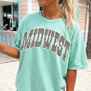 Puede incluir: Camiseta verde claro con la palabra "MIDWEST" en un diseño de estampado de leopardo. La camiseta tiene un corte holgado. La persona lleva gafas de sol y joyas. El fondo es una playa.