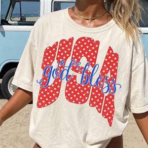 Puede incluir: Camiseta color crema con la frase "USA God Bless" en letras de lunares rojos y escritura cursiva azul. La camiseta tiene un corte holgado, ideal para uso casual. Una furgoneta azul claro se ve en el fondo.