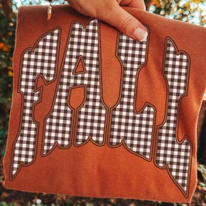 Pode incluir: Uma t-shirt de cor laranja queimado com a palavra FALL em letras grandes e estilizadas. As letras têm um padrão de xadrez vichy preto e branco e são contornadas por uma fina borda castanha. O design está impresso na frente do tecido.