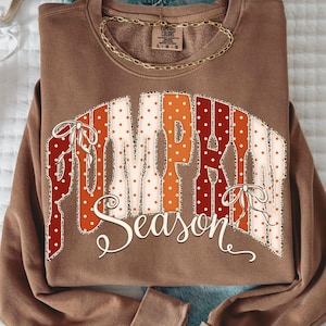 May include: A brown crewneck sweatshirt featuring the text PUMPKIN SEASON in a large, arched graphic. The letters are filled with a pattern of white polka dots on orange and red backgrounds, accented with leopard print edges and white bows.