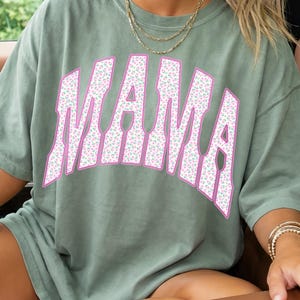 Peut inclure: T-shirt vert olive avec le mot "MAMA" en grandes lettres roses &agrave; motifs floraux. La personne assise dans une voiture porte le t-shirt. La personne porte des lunettes de soleil et des bijoux.
