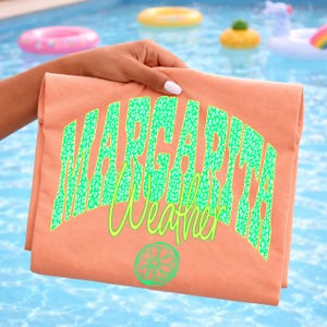 May include: A folded peach-colored t-shirt with the words "Margarita Weather" in green leopard print lettering and a lime graphic. The shirt is held up against a blue pool background with inflatable pool toys.