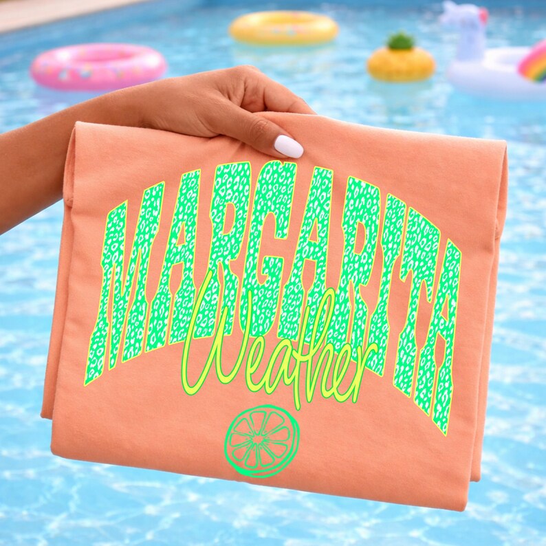 May include: A folded peach-colored t-shirt with the words "Margarita Weather" in green leopard print lettering and a lime graphic. The shirt is held up against a blue pool background with inflatable pool toys.