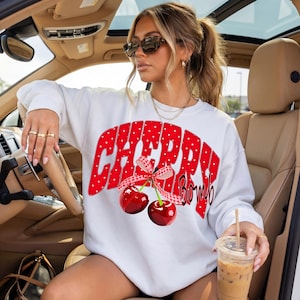 May include: White sweatshirt with the text "CHERRY BOMB" in red, polka-dotted letters. Below the text are two cherries tied with a red and white checkered ribbon. The sweatshirt is a casual, everyday garment.