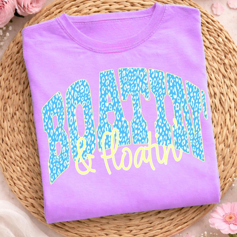 May include: A lilac t-shirt featuring the text "BOATIN & Floatin" in a blue leopard print design and yellow script. The shirt is folded on a woven mat, with pink flowers in the background.