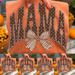 May include: An orange t-shirt featuring the word MAMA in large brown letters with white polka dots. A brown and white gingham patterned bow is centered below the text. The design is printed on the front of the fabric in a bold, western-style font.