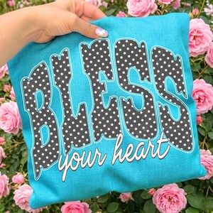 May include: A turquoise pillow with the words "BLESS your heart" in a playful font. The letters are filled with a black and white polka dot pattern. The pillow is surrounded by pink roses.