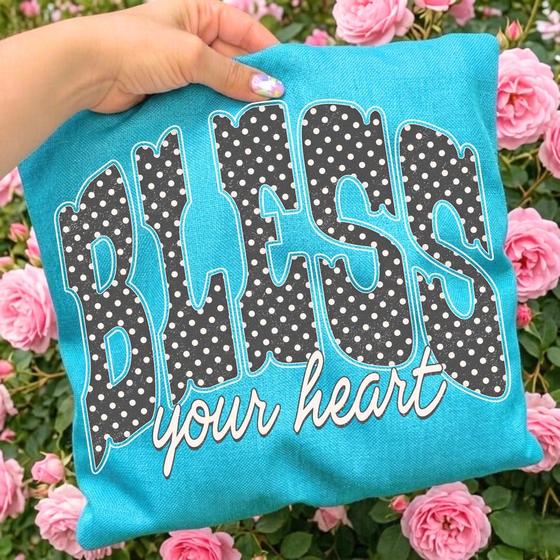 May include: A turquoise pillow with the words "BLESS your heart" in a playful font. The letters are filled with a black and white polka dot pattern. The pillow is surrounded by pink roses.