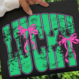 May include: Black tote bag with the word "LUCKY" in large, distressed green letters outlined in pink. Two pink bows are placed on the letters. The bag is held by a person.