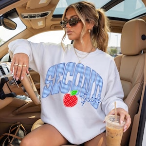 May include: White sweatshirt with the words "SECOND Grade" in blue gingham print, a red apple graphic, and the word "Grade" in blue script. The sweatshirt is worn by a person in a car.