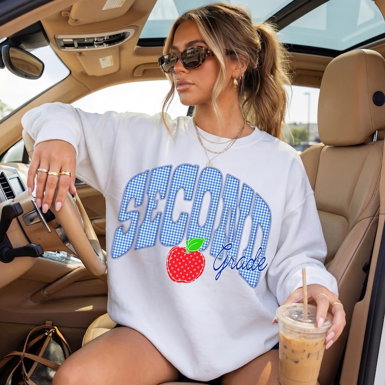 May include: White sweatshirt with the words "SECOND Grade" in blue gingham print, a red apple graphic, and the word "Grade" in blue script. The sweatshirt is worn by a person in a car.