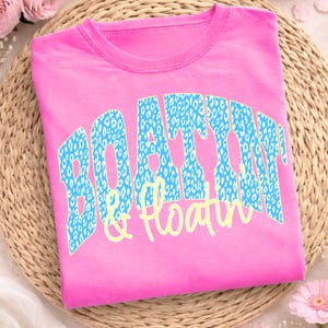 May include: A vibrant pink t-shirt featuring the text "BOATTIN' & Floatin'" in a blue leopard print design. The text is arched across the front of the shirt. The shirt is folded neatly on a woven surface.