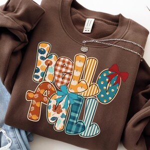 May include: A brown sweatshirt with the words "Hello Fall" in a colorful, patterned design. The letters are decorated with pumpkins, leaves, and plaid patterns. A silver necklace with a small charm is draped across the sweatshirt.