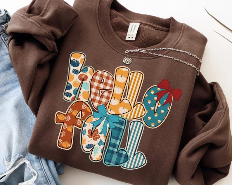 May include: A brown sweatshirt with the words "Hello Fall" in a colorful, patterned design. The letters are decorated with pumpkins, leaves, and plaid patterns. A silver necklace with a small charm is draped across the sweatshirt.