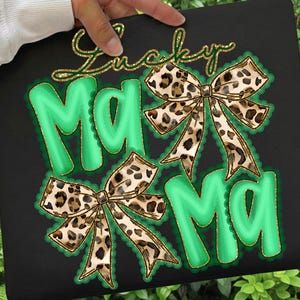 May include: Black graphic tee with the words "Lucky Mama" in gold glitter script. The word "Mama" is in bright green with a gold trim and two leopard print bows. The design is on a black background.