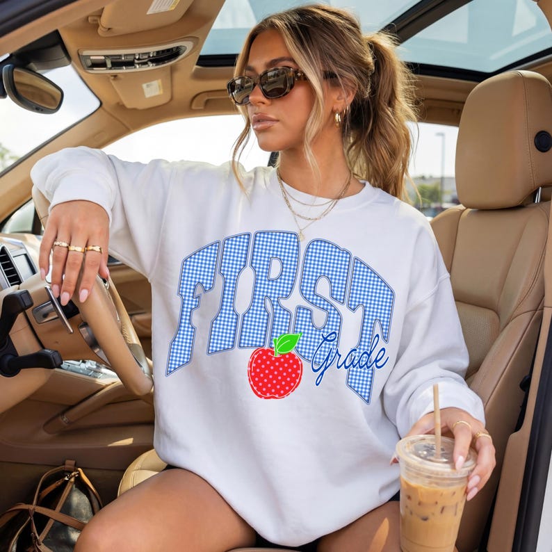 May include: White sweatshirt with the words "FIRST Grade" in blue gingham print. A red apple graphic with a green leaf is below the text. The sweatshirt is a casual style.