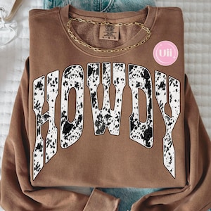 Könnte beinhalten: Ein braunes Sweatshirt mit Rundhalsausschnitt und dem Wort HOWDY in großen, geschwungenen Blockbuchstaben. Die Buchstaben haben ein weiß-schwarzes Kuhfellmuster. Eine goldene Kette liegt am Kragen und ein kleines rosa kreisförmiges Logo mit dem Text uii ist zu sehen.