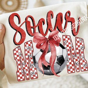 May include: Cream-colored t-shirt with the words "Soccer Mom" in red script and block letters. A soccer ball with a red bow is in the center. The letters "M" are decorated with a red and white checkered pattern.