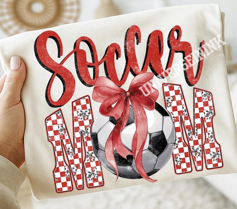 May include: Cream-colored t-shirt with the words "Soccer Mom" in red script and block letters. A soccer ball with a red bow is in the center. The letters "M" are decorated with a red and white checkered pattern.
