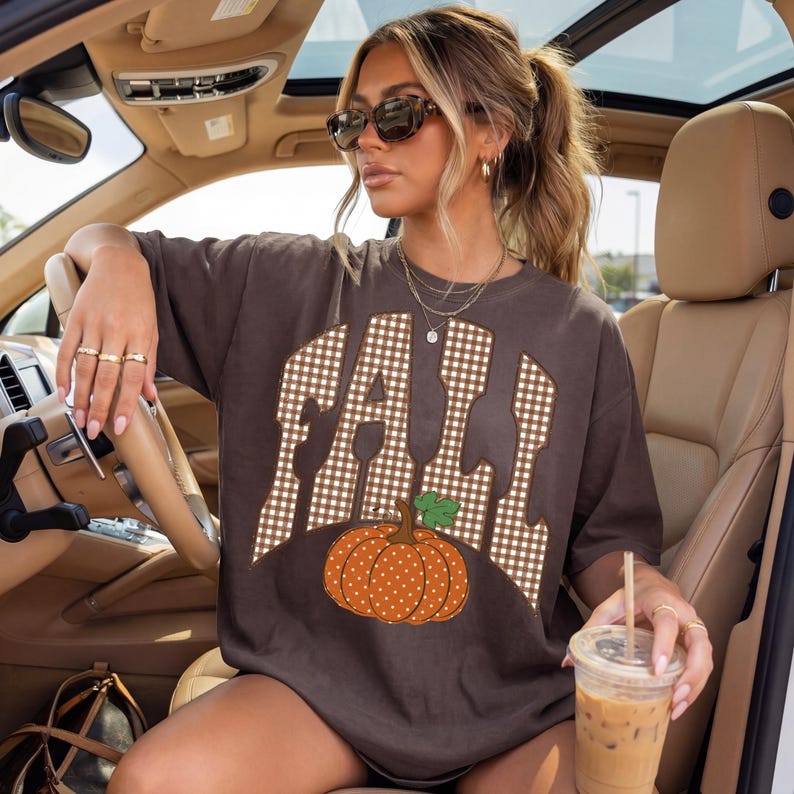 Peut inclure: Un t-shirt &agrave; manches courtes marron fonc&eacute; avec le mot FALL &eacute;crit dans un motif vichy blanc et marron. Sous le texte se trouve un graphique repr&eacute;sentant une citrouille orange &agrave; pois blancs avec une tige verte. Col rond et coupe ample.