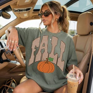 Peut inclure: Un t-shirt &agrave; manches courtes vert olive avec le mot FALL &eacute;crit dans un motif &agrave; carreaux vichy marron et blanc. Sous le texte se trouve un graphique repr&eacute;sentant une citrouille orange &agrave; pois blancs avec une tige verte. Col rond.