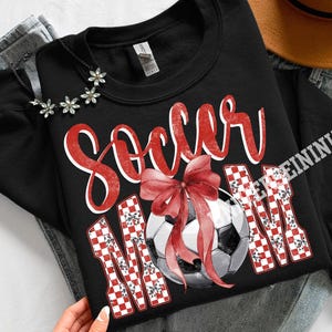 May include: Black sweatshirt with the words "Soccer Mom" in red script and checkered letters. A soccer ball with a red bow is in the center. The sweatshirt is on a denim background.