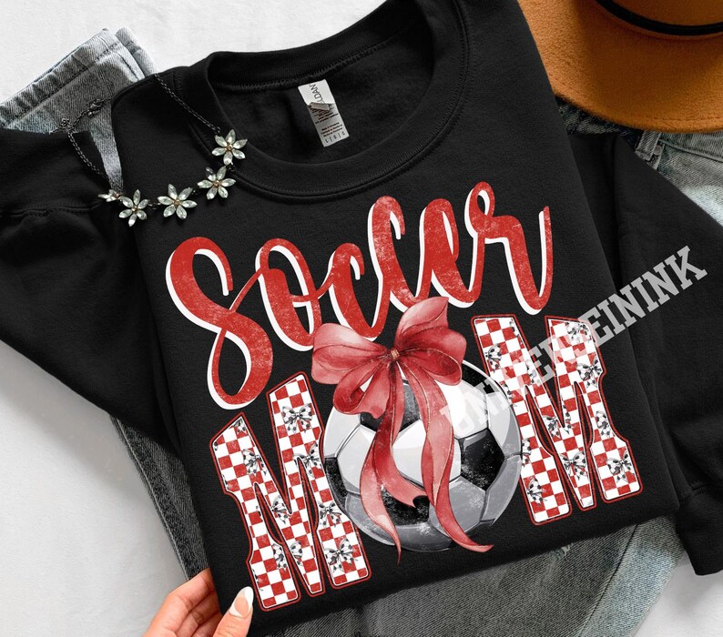 May include: Black sweatshirt with the words "Soccer Mom" in red script and checkered letters. A soccer ball with a red bow is in the center. The sweatshirt is on a denim background.