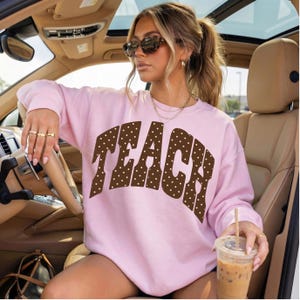 Könnte beinhalten: Ein hellrosa langärmeliges Sweatshirt mit Rundhalsausschnitt und dem Wort TEACH in großen, braunen Blockbuchstaben mit weißem Punktemuster auf der Brust. Das Kleidungsstück hat eine lockere Passform mit gerippten Bündchen und einem gerippten Halsausschnitt.