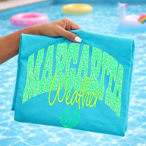 May include: A folded turquoise t-shirt with the words "Margarita Weather" in a neon green leopard print font. A lime slice graphic is below the text. The shirt is held up by a hand in front of a swimming pool.