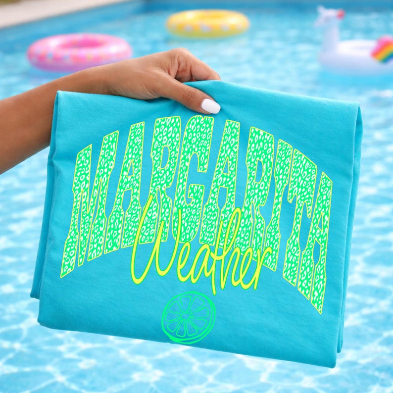 May include: A folded turquoise t-shirt with the words "Margarita Weather" in a neon green leopard print font. A lime slice graphic is below the text. The shirt is held up by a hand in front of a swimming pool.