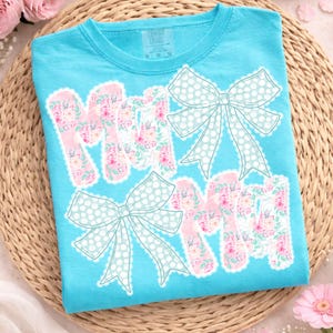 May include: A turquoise t-shirt featuring the word "MOM" in pink floral letters with white scalloped edges. Light blue polka dot bows are above and below the word. The shirt is made of a soft material.