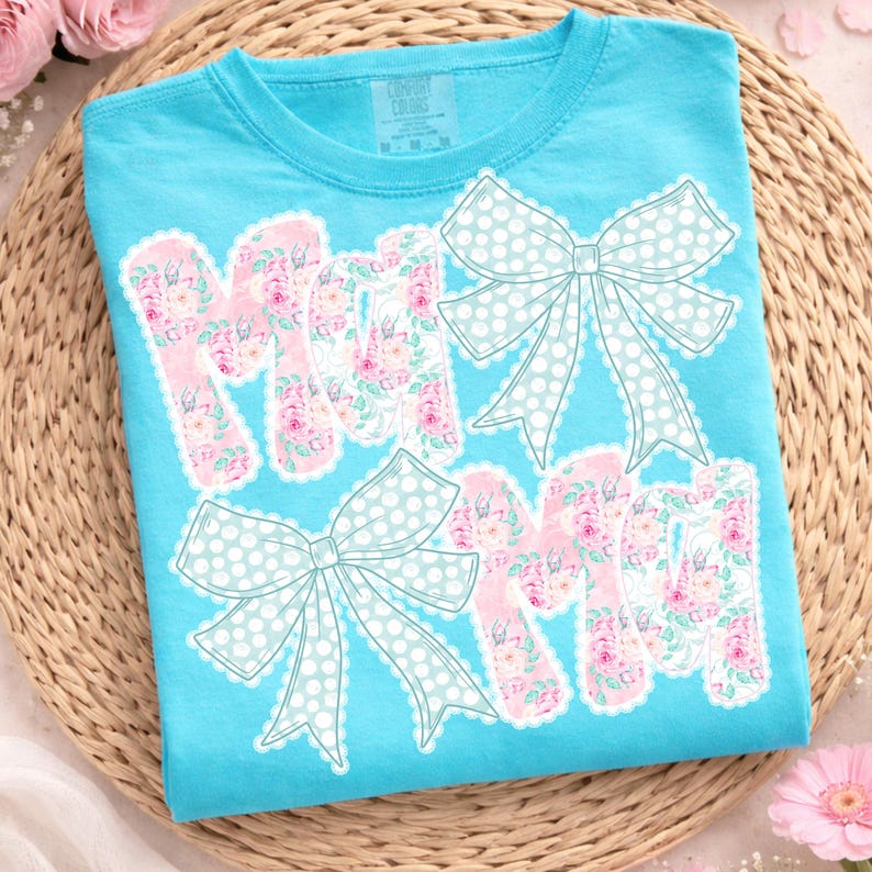 May include: A turquoise t-shirt featuring the word "MOM" in pink floral letters with white scalloped edges. Light blue polka dot bows are above and below the word. The shirt is made of a soft material.