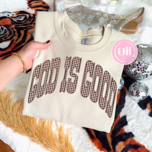 May include: A cream-colored crewneck sweatshirt featuring the text "GOD IS GOOD" printed in a brown and white gingham pattern. The lettering has a curved, bold font style. The garment has a ribbed collar and a standard long-sleeve construction.